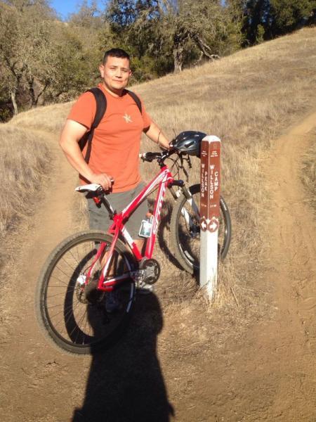 A person standing next to a mountain bike at a trail marker on a dirt path, surrounded by dry grass and trees in the background. The individual is wearing a casual orange t-shirt and shorts, smiling while holding onto the bike. Henry W. Coe State Park mountain bike trail.