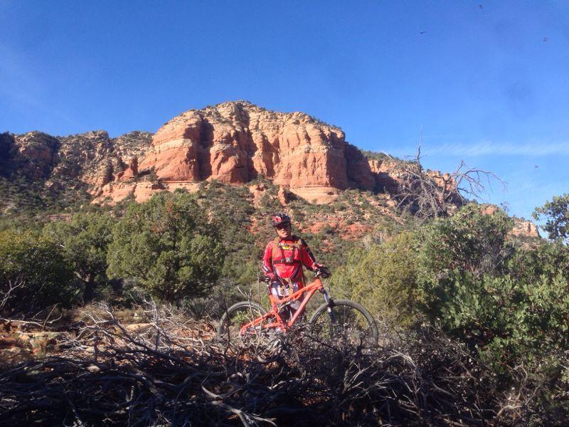 A mountain biker in a red jersey stands next to an orange bike in a desert landscape, with striking red rock formations in the background and green shrubs surrounding the area under a clear blue sky. Hiline mountain bike trail.