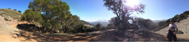 A panoramic view of a scenic outdoor landscape featuring rolling hills, a large green tree, and a person standing in the foreground. The sun shines brightly in the sky, with a clear view of distant mountains in the background. Pathways wind through the terrain, suggesting a hiking or biking trail. Camp Tamarancho mountain bike trail.
