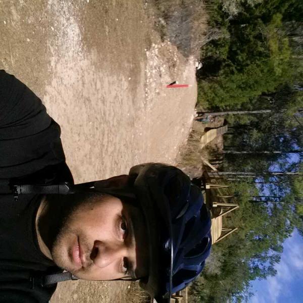 A person wearing a black cycling helmet, standing on a dirt path in a wooded area. The background features trees and an unfinished wooden structure, with a red marker visible on the path. The sky is clear and blue. Santos mountain bike trail.