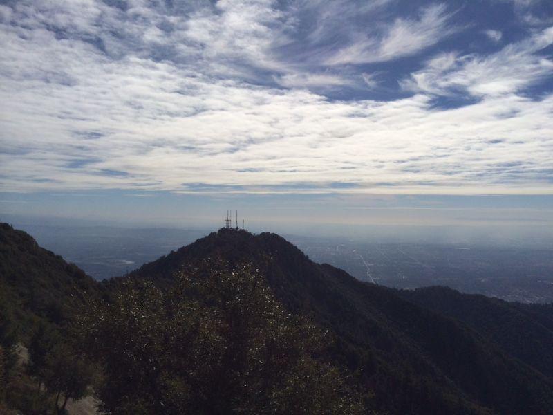 A panoramic view from a mountain peak overlooking a vast landscape. Wispy clouds fill the sky above, while antennas are visible on the summit. Lush greenery covers the mountain slopes, contrasting with the distant cityscape beneath a haze. Mount Wilson Trail mountain bike trail.