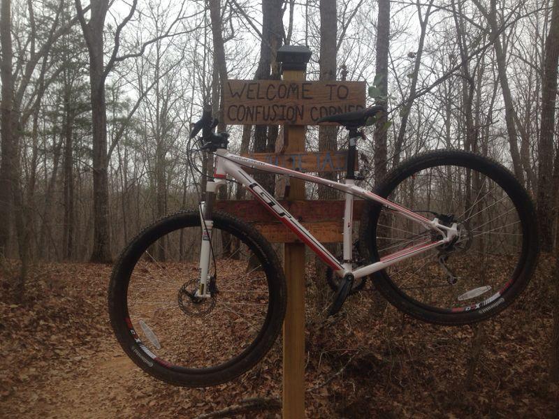 A mountain bike hanging on a wooden sign that reads "Welcome to Confusion Corner," surrounded by bare trees and leaf-covered ground on a trail. Chicopee Woods mountain bike trail.