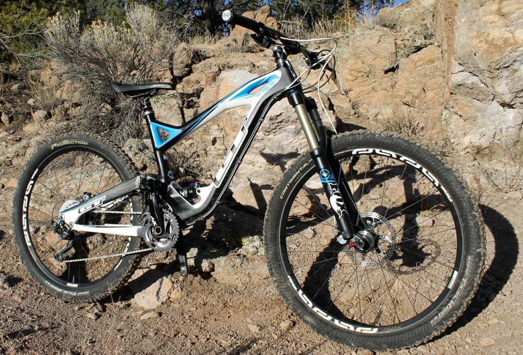 GT Force Carbon Pro: A mountain bike leaning against a rocky surface, featuring a sleek design with a blue and white frame, thick tires, and visible gears and brakes. The background includes dry vegetation and rocky terrain, indicating an outdoor setting.