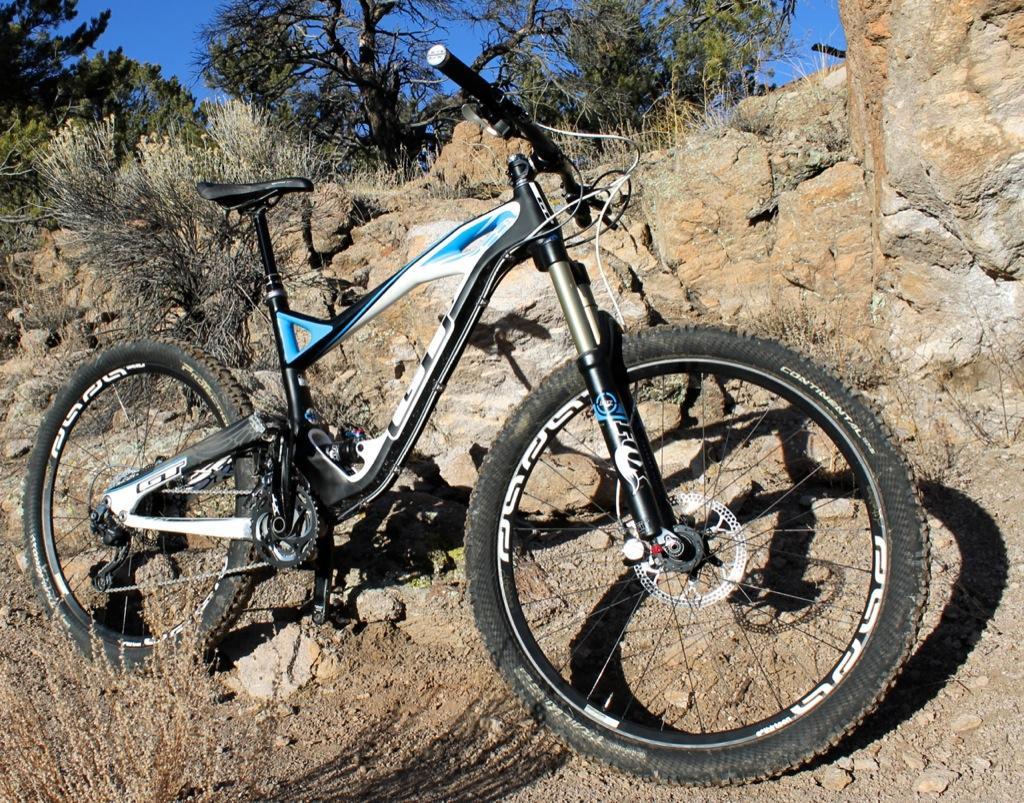 GT Force Carbon Pro: A mountain bike positioned on rocky terrain, featuring a black and white frame with blue accents. The bike is equipped with thick, knobby tires suitable for off-road riding. In the background, there are shrubs and trees under a clear blue sky, showcasing an outdoor setting ideal for cycling adventures.