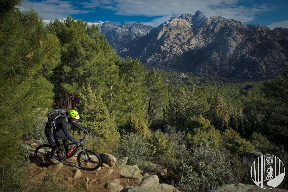A mountain biker navigates a rocky trail through a dense forest, with snow-capped mountains in the background under a partly cloudy sky. The biker wears a bright yellow helmet and rides a black mountain bike, surrounded by lush greenery and rugged terrain. La Pedriza PR-16 mountain bike trail.