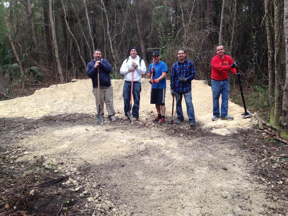 Five men standing in a wooded area, each holding a gardening or landscaping tool, with a small mound of dirt and debris in the background. They are positioned in front of a newly cleared section of ground, surrounded by trees and greenery. Tillie Fowler Regional Park mountain bike trail.