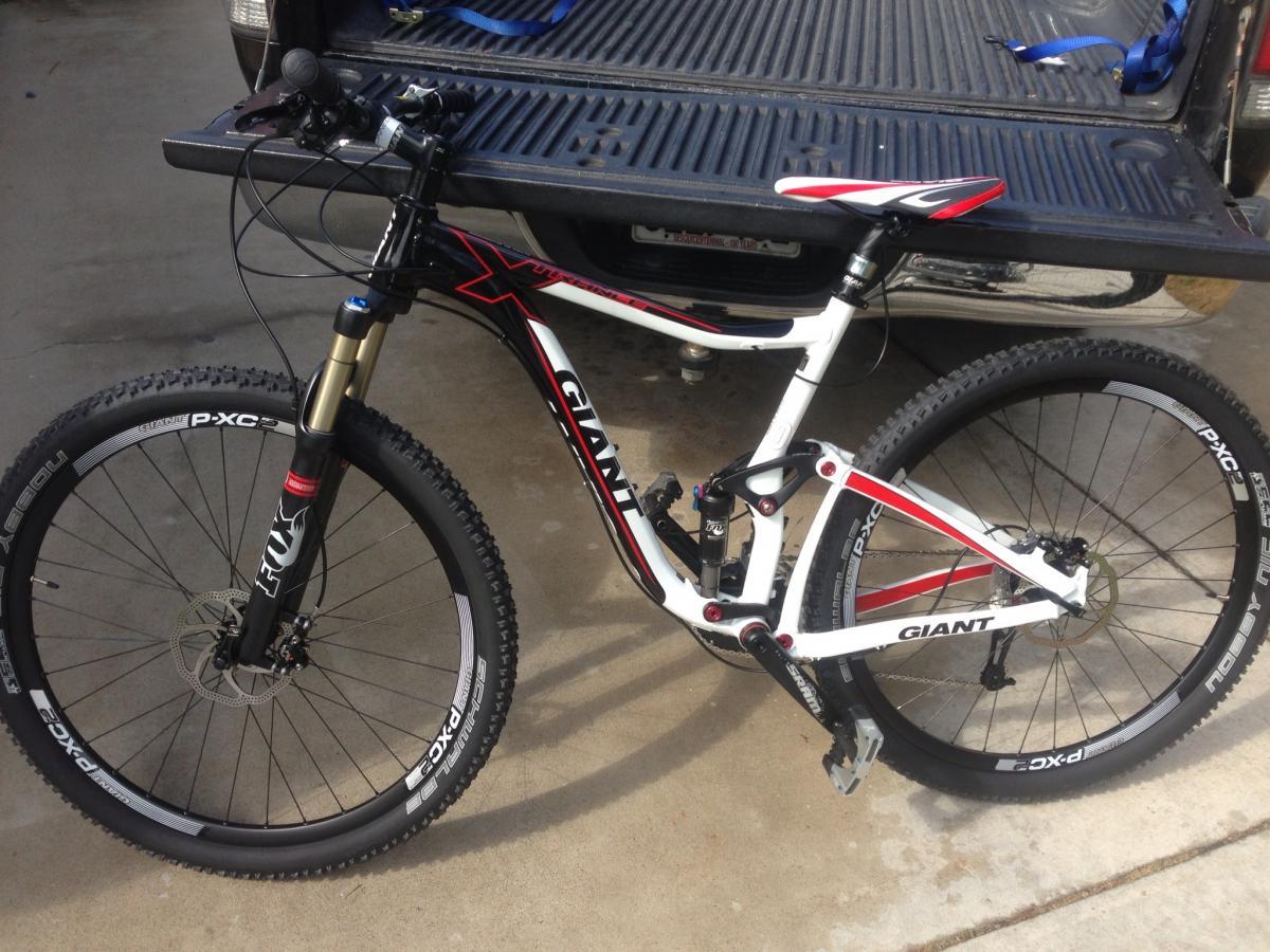 Giant Trance X 29er: A close-up view of a white and black mountain bike, featuring a prominent "Giant" logo. The bike has a front suspension fork, disc brakes, and knobby tires designed for off-road terrain. In the background, there's a truck bed with a textured surface. The setting appears to be an outdoor concrete area.