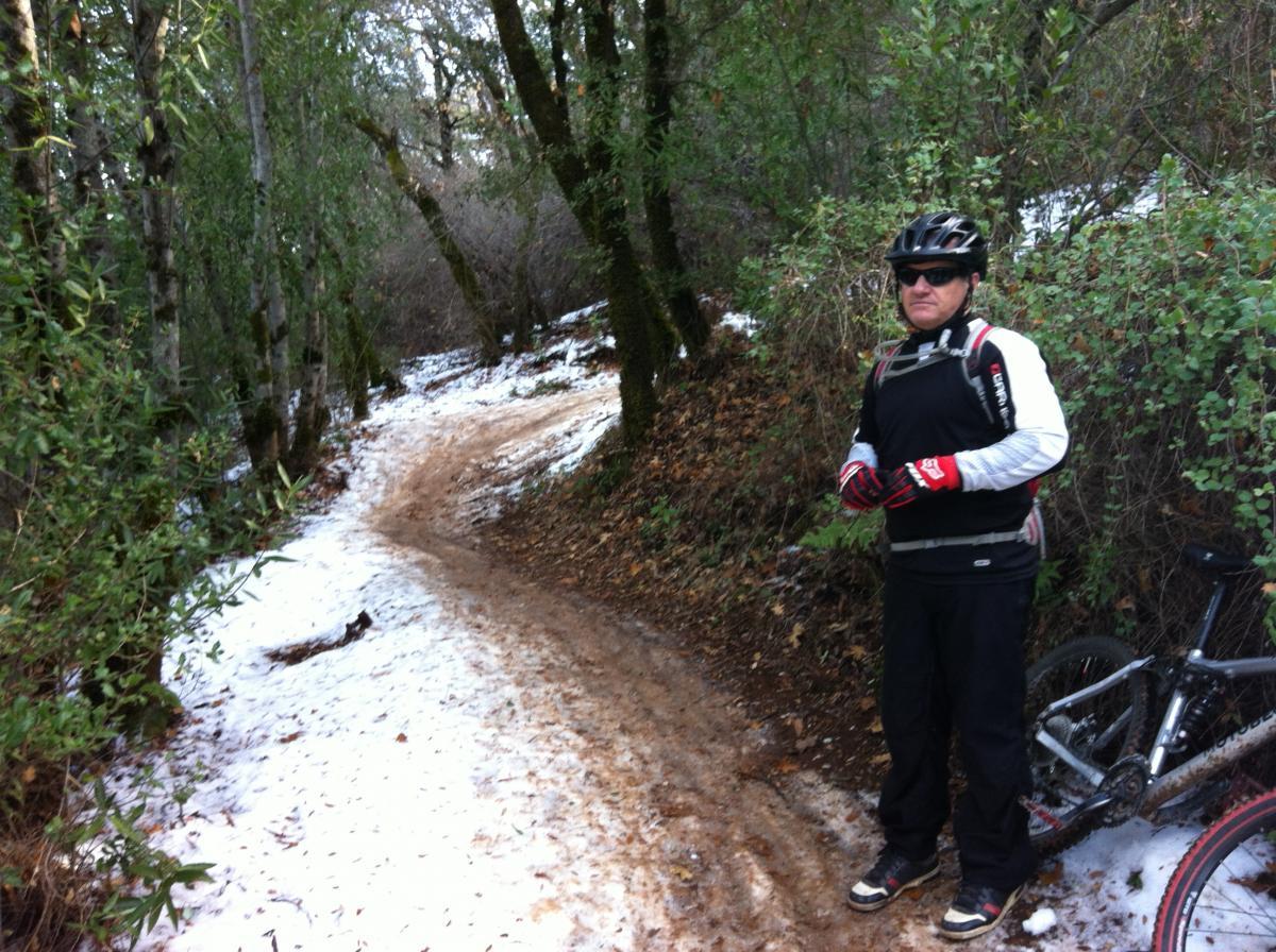 A mountain biker wearing a helmet and protective gear stands beside a snowy, dirt trail surrounded by trees. A bicycle is parked near him, and the trail curves into the distance. The scene depicts a mix of snow and earthy terrain in a wooded area. Clementine / Forresthill Connector Trail mountain bike trail.