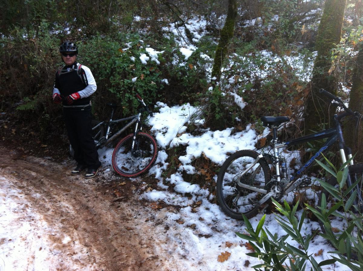 A mountain biker standing next to two bikes on a snowy trail, surrounded by greenery and fallen leaves. The biker is wearing a helmet, sunglasses, and gloves, and is dressed in black winter cycling attire. Snow is visible on the ground alongside a muddy path, indicating a mixed weather condition. Clementine / Forresthill Connector Trail mountain bike trail.