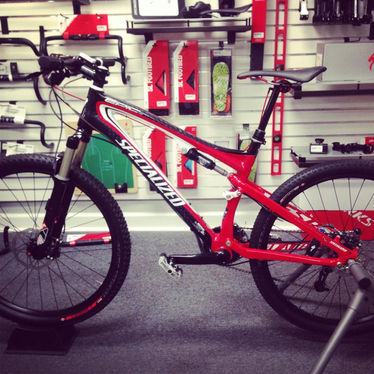 Specialized EPIC COMP CARBON: A red and black mountain bike displayed in a store, featuring a lightweight frame, front suspension, and rugged tires. Various bike accessories and parts are visible in the background on shelves.