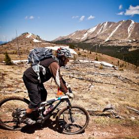A mountain biker riding on a dirt trail in a mountainous landscape, surrounded by snow-capped peaks and blue skies. The cyclist is wearing a helmet and a backpack, leaning slightly forward as they navigate the terrain.