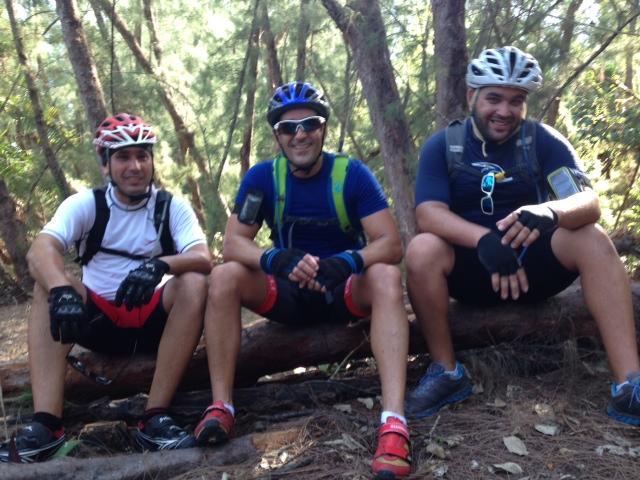 Three men sitting on a log in a forested area, wearing cycling gear and helmets. They are taking a break from biking, smiling at the camera. The background features tall trees and a natural, green setting. Oleta River State Park mountain bike trail.