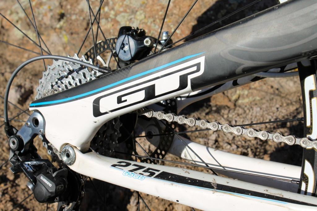 GT Force Carbon Pro: Close-up view of the rear section of a GT mountain bike, featuring the bike's distinctive logo, gear mechanism, and chain. The background shows rocky terrain, indicating a rugged outdoor setting.