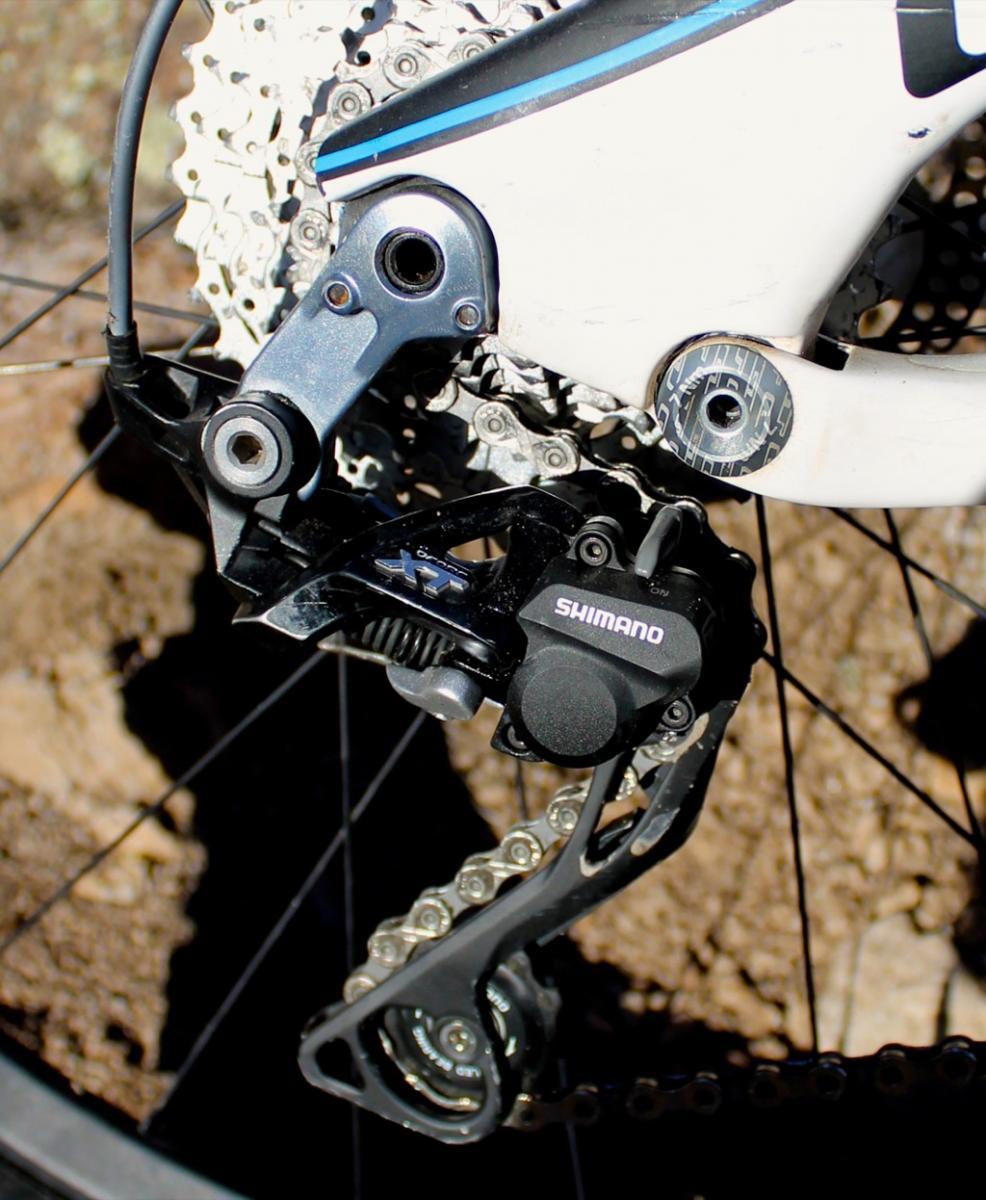 GT Force Carbon Pro: A close-up of a Shimano bicycle rear derailleur mounted on a bike frame, with a visible gear cassette and chain in the background. The image shows details of the derailleur's design, including its mechanical components and connections, set against a natural, earthy backdrop.