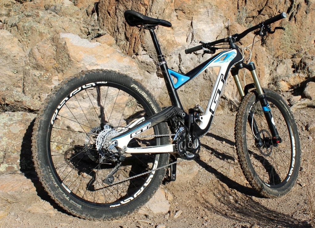GT Force Carbon Pro: A mountain bike with a black and white frame accented by blue details, resting against a rocky background. The bike features large, rugged tires suitable for off-road terrain and displays a rear gear system, showcasing its design for mountainous rides.