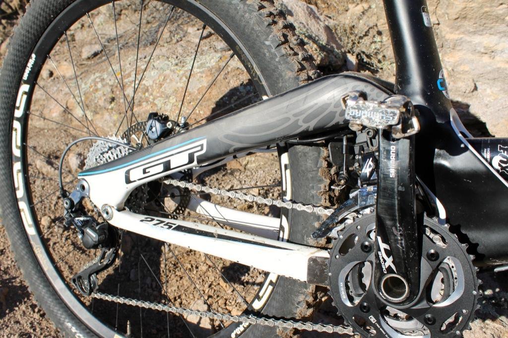 GT Force Carbon Pro: A close-up view of a GT mountain bike's drivetrain, featuring the rear derailleur, chain, and crankset. The bike is positioned on rocky terrain, showcasing its rugged design and components. The frame has a sleek black and gray color scheme, highlighting the model name and specifications.