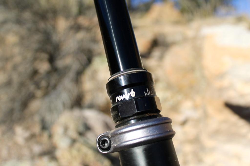 GT Force Carbon Pro: Close-up image of a bicycle dropper post mechanism, featuring a black anodized surface with a visible adjustment knob and markings for height settings. The background shows blurred natural terrain with rocks and vegetation.