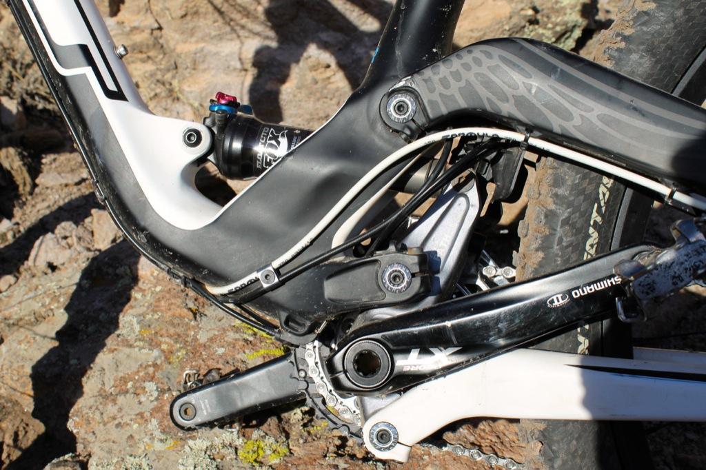 GT Force Carbon Pro: Close-up view of a mountain bike's bottom bracket area, showcasing the frame's detailed design, suspension components, and drivetrain, with sunlight highlighting the textures and colors against a rocky background.