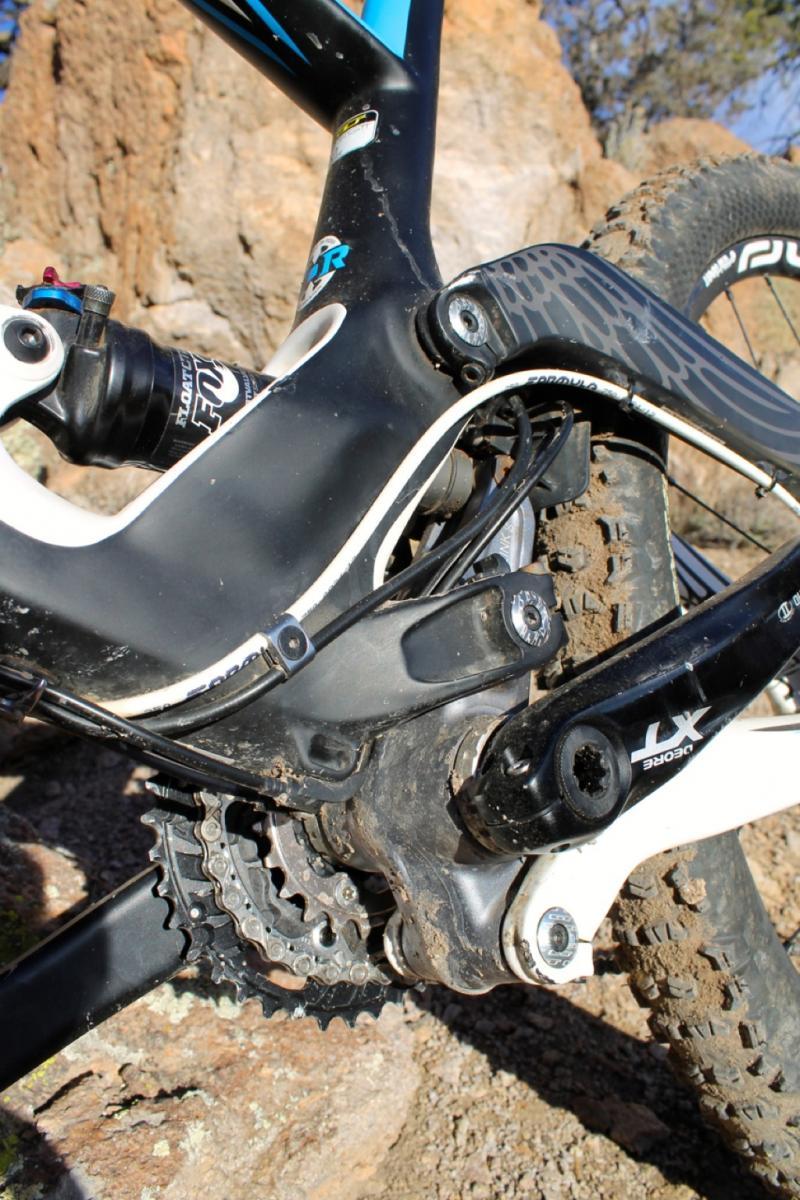 GT Force Carbon Pro: Close-up view of a mountain bike's rear triangle, showcasing the suspension system, chainrings, and drivetrain components. The frame features a black and blue design, with visible dirt and grime indicating recent use on rugged terrain. The background includes rocky ground and sparse vegetation.