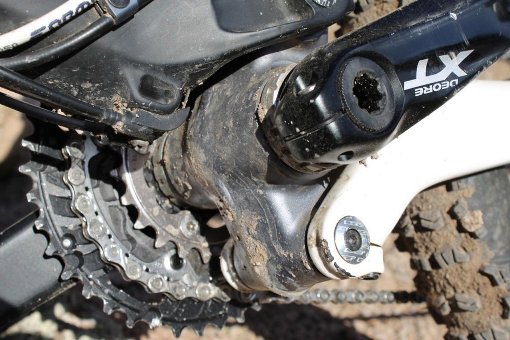 GT Force Carbon Pro: Close-up view of a bike's bottom bracket and crankset, showing a muddy and worn drivetrain with chainrings, pedals, and cables, indicating use in off-road conditions.
