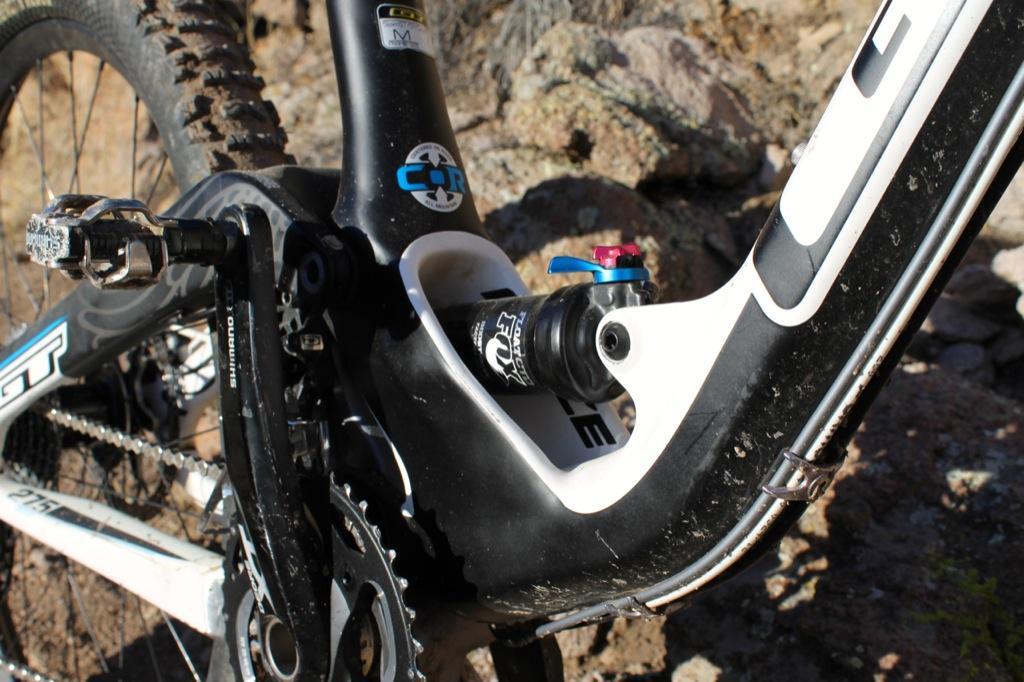 GT Force Carbon Pro: Close-up shot of a mountain bike's bottom bracket and drivetrain, featuring a Shimano pedal, chainring, and a visible shock component. The bike has a black and white frame with some dirt and mud, indicating recent outdoor use. Background shows rocky terrain.