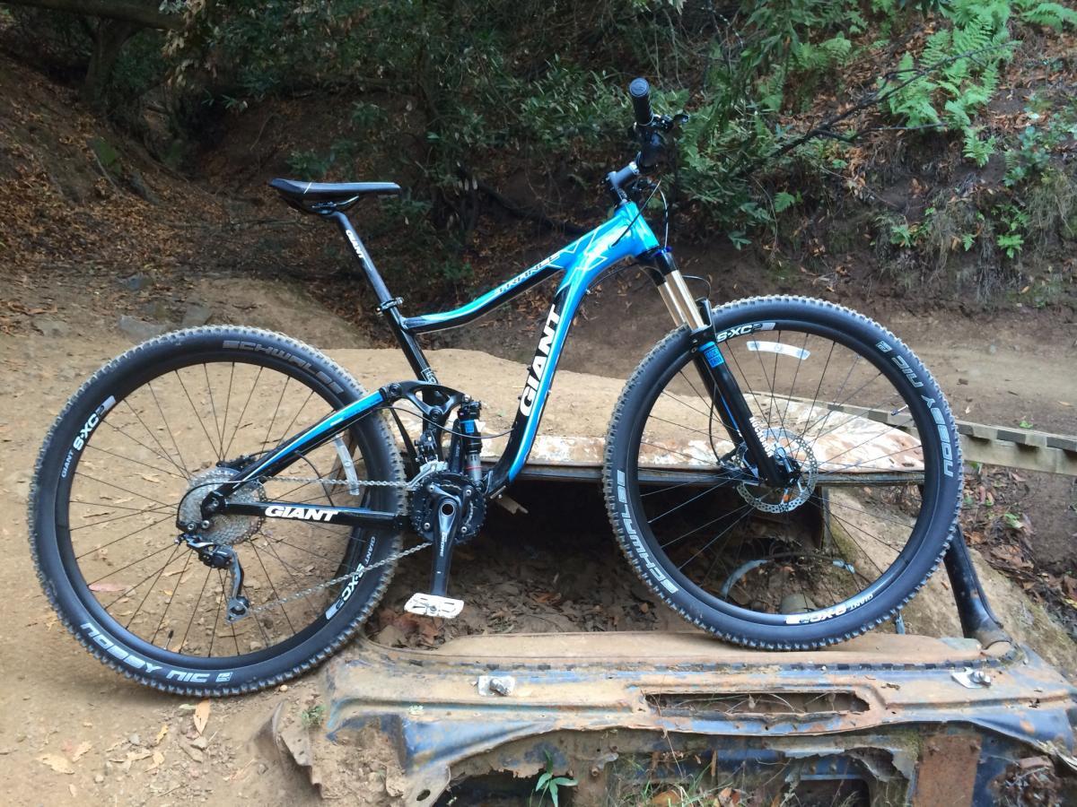 Giant Trance X 29er: A blue and black mountain bike resting on an old, rusted vehicle chassis in a wooded area. The bike features wide tires, a front suspension fork, and multiple gears, surrounded by earthy tones and foliage in the background.