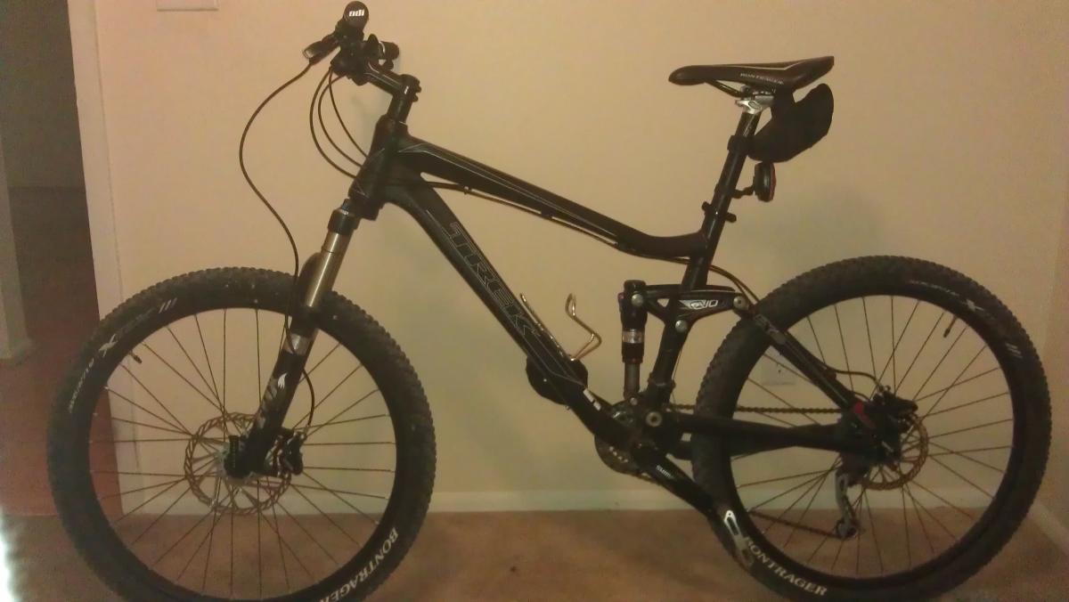 Trek Fuel EX 8: A black mountain bike with front suspension, thick tires, and disc brakes, positioned upright against a wall. The bike features a streamlined frame and is equipped with a saddle bag. The background includes a neutral-colored wall and floor.