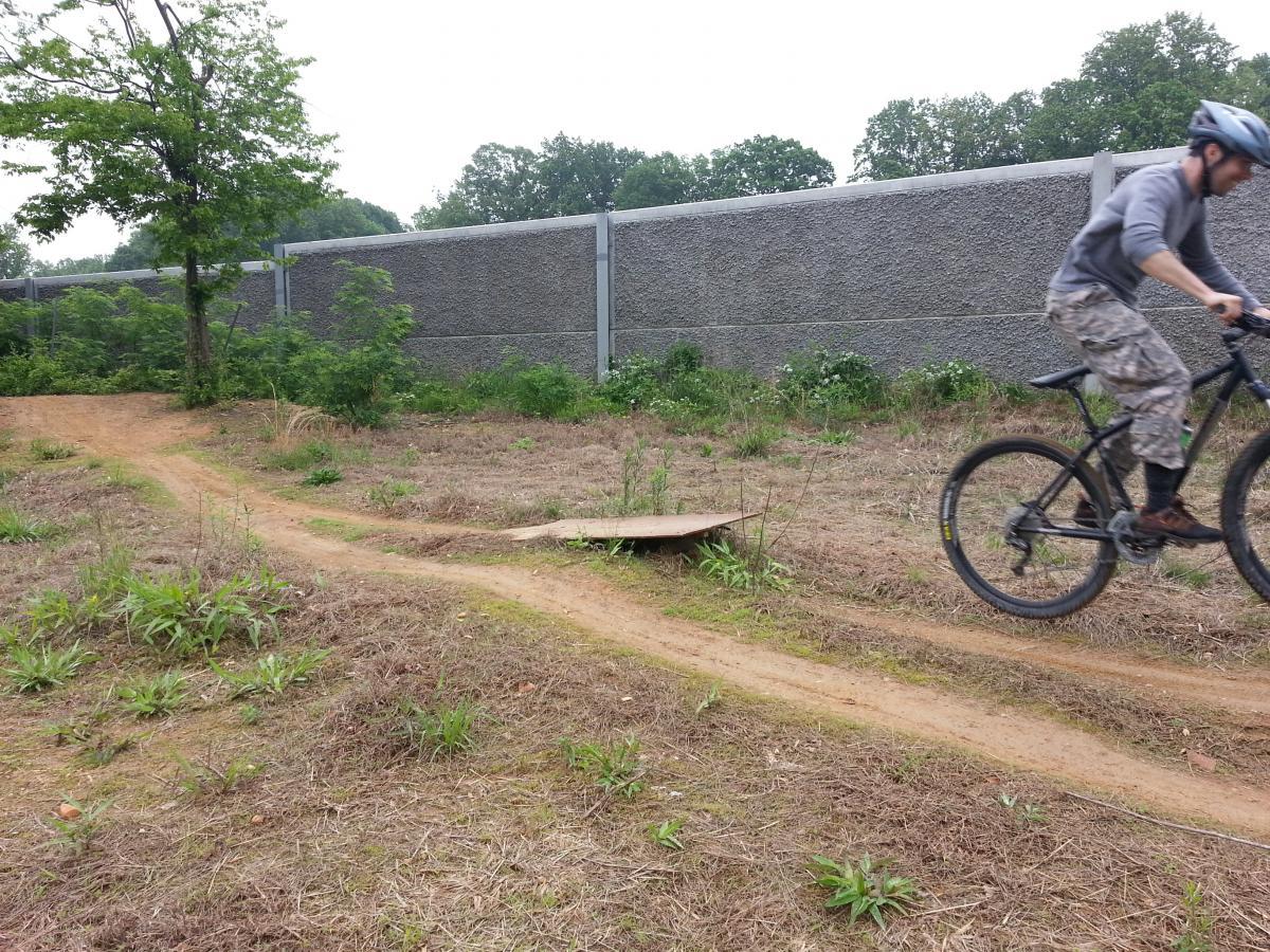 A cyclist in a gray helmet and casual clothing is mid-jump over a small wooden ramp on a dirt path surrounded by greenery. In the background, a gray wall and dense foliage are visible under a cloudy sky. Wakefield mountain bike trail.