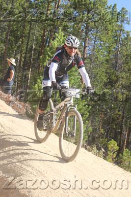 Trek Superfly 100 Elite carbon: A mountain biker riding on a dirt trail through a forested area, wearing a helmet and cycling gear. In the background, there are trees and a person observing the ride.