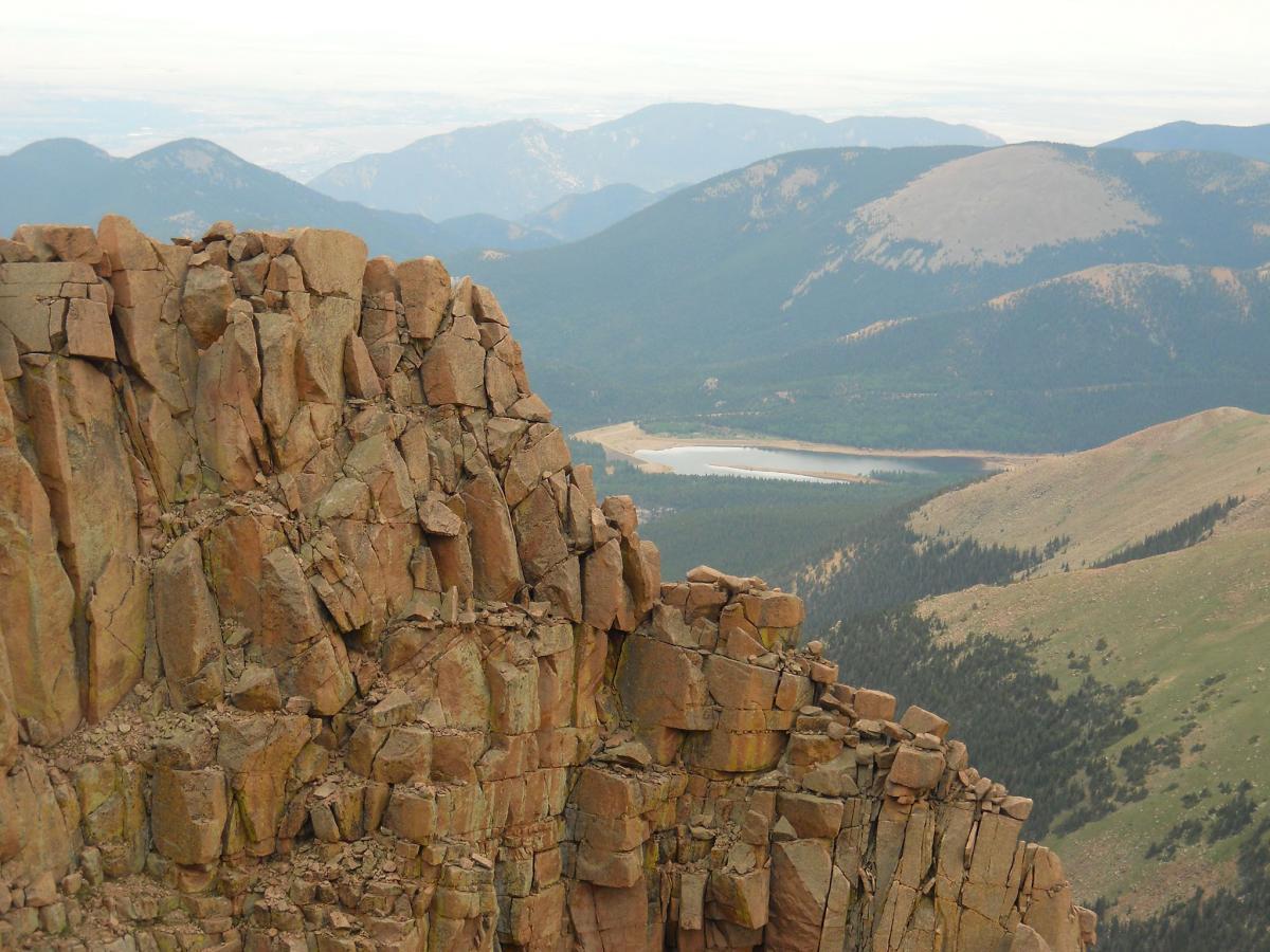 A rocky cliff edge overlooking a vast mountainous landscape, with layers of jagged rocks in the foreground. The background features rolling hills and distant mountains, along with a calm lake nestled in the valley below. The atmosphere is slightly cloudy, providing a soft, diffused light across the scene. Barr Trail / Pikes Peak mountain bike trail.