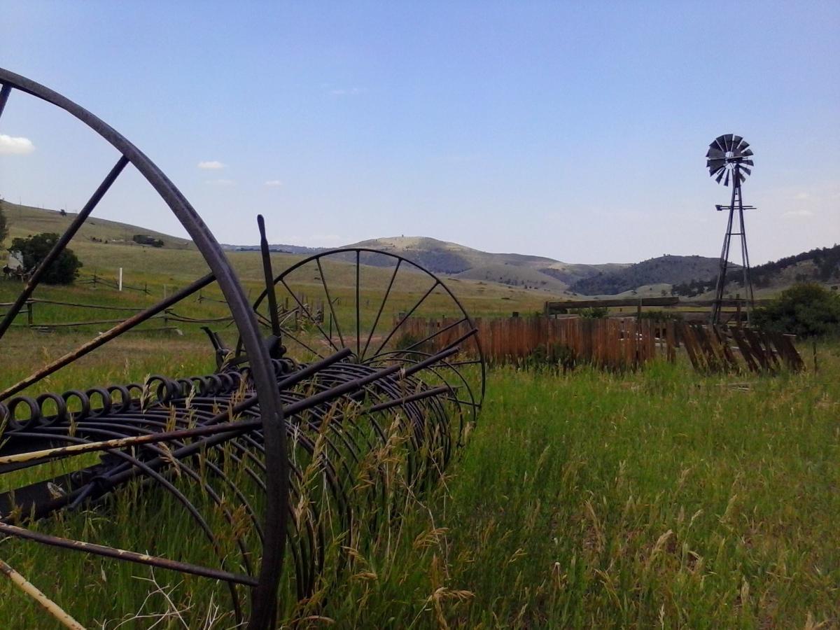 A scenic view of a rural landscape featuring vintage agricultural equipment, including a large metal hay rake, set against rolling green hills. In the background, a classic windmill stands tall, surrounded by lush grass and a wooden fence, under a clear blue sky with a few clouds. Centennial Cone Park mountain bike trail.