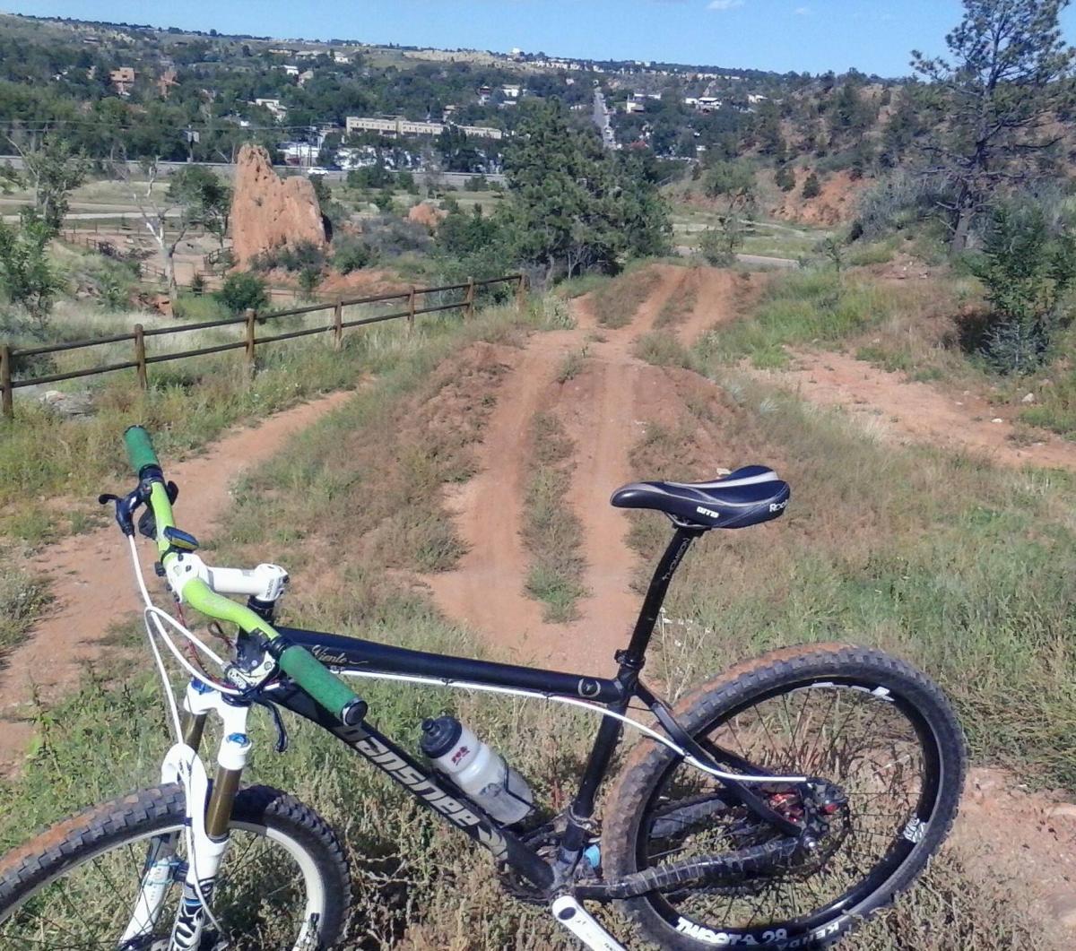 A mountain bike is parked on a dirt trail, surrounded by grass and brush. In the background, a rocky outcrop and a small town are visible on a hillside. The area is sunny with a clear sky, suggesting an ideal setting for outdoor adventures. Red Rock Canyon mountain bike trail.