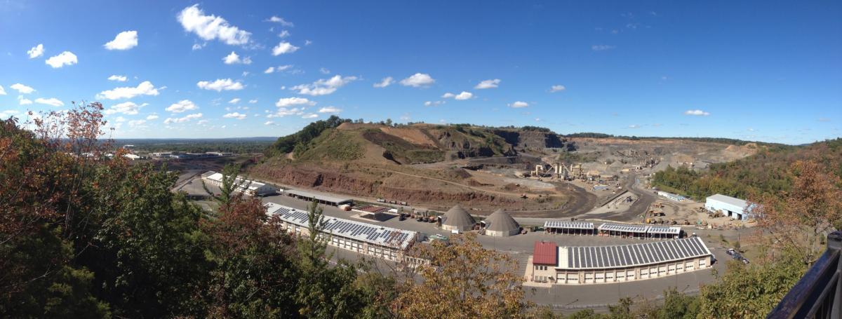 A panoramic view of a mining site, featuring large excavated areas on the hills, machinery, and storage facilities. The landscape is dotted with trees showing autumn colors, under a partly cloudy sky. Chimney Rock mountain bike trail.