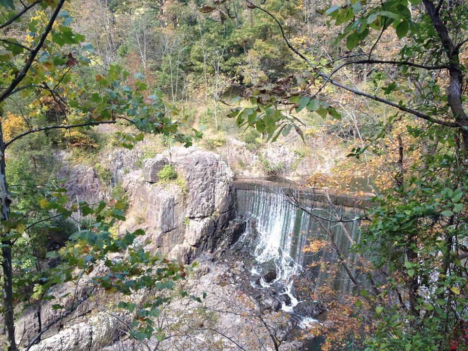 A scenic view of a waterfall cascading over a rocky ledge, surrounded by lush green trees and autumn foliage. The image captures the natural beauty of the landscape, with vibrant colors highlighting the change of seasons. Chimney Rock mountain bike trail.