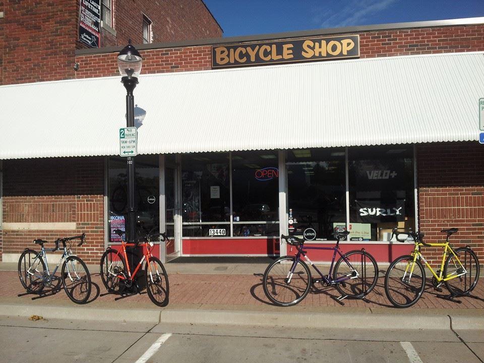 A bicycle shop with a brick exterior featuring a white awning. The shop displays a large "BICYCLE SHOP" sign above the entrance, which has large windows showcasing various bicycles and an "OPEN" neon sign. Five bicycles in different colors (blue, red, black, yellow, and purple) are parked outside on the sidewalk. A streetlamp and parking sign can also be seen in the image.