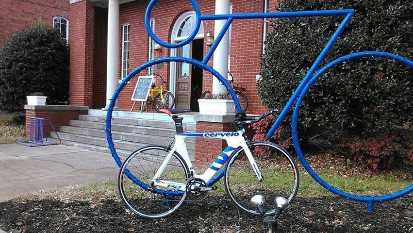 White and blue Cervelo road bike leaning against a large blue bicycle sculpture in front of a brick building, with a small decorative bike and steps leading to the entrance.