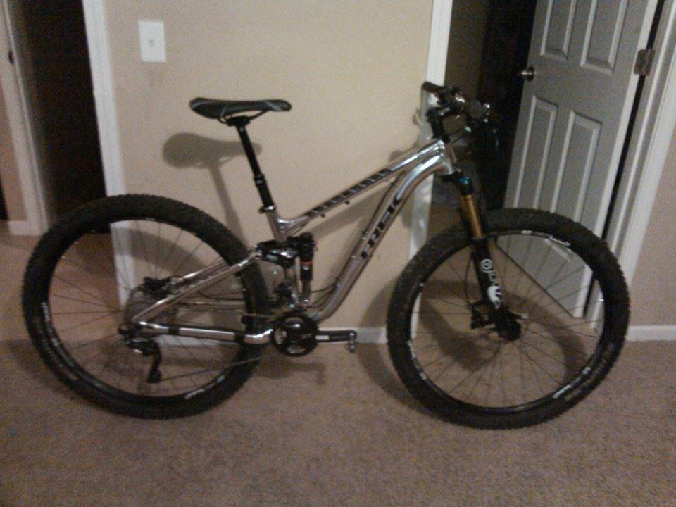 Trek Fuel EX 9 29: A silver mountain bike with thick tires is positioned inside a room. The bike features a black seat and handlebars, and it rests on a carpeted floor with a door visible in the background.