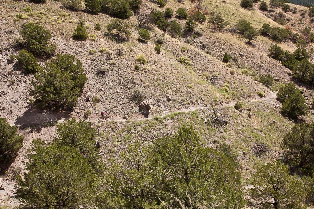 A scenic view of a mountainous landscape featuring a winding dirt path. A person rides a mountain bike along the trail, surrounded by patches of green vegetation and trees on rocky terrain. The scene captures the natural beauty of the outdoors. Arkansas Hills mountain bike trail.