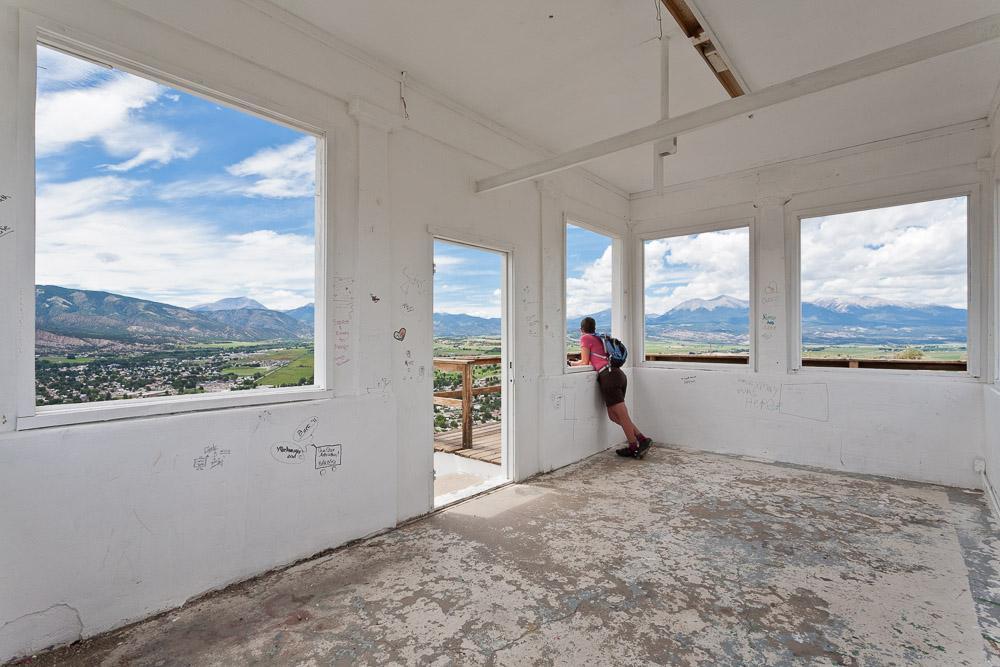 A person stands indoors, gazing out of large windows that frame a picturesque view of mountains and a valley below. The walls are bare and show signs of wear, while the floor appears worn. Bright clouds drift across a blue sky, enhancing the scenic landscape outside. Arkansas Hills mountain bike trail.