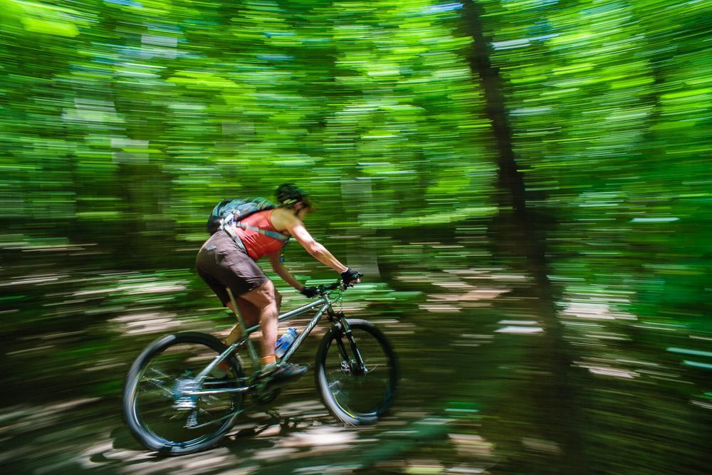 A person biking rapidly through a lush green forest, captured with a motion blur effect to convey speed and movement. Hatchery Creek Trail mountain bike trail.