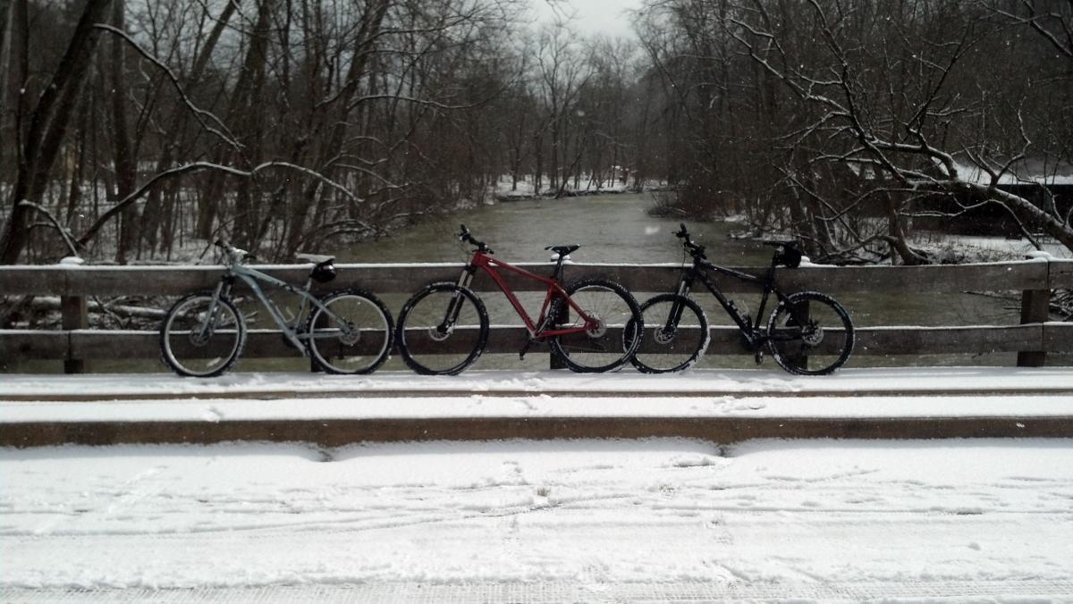 Trek Stache: Three bicycles are parked on a snow-covered bridge overlooking a winding river, surrounded by bare trees. The scene captures a serene winter landscape with snow gently falling, creating a tranquil atmosphere.
