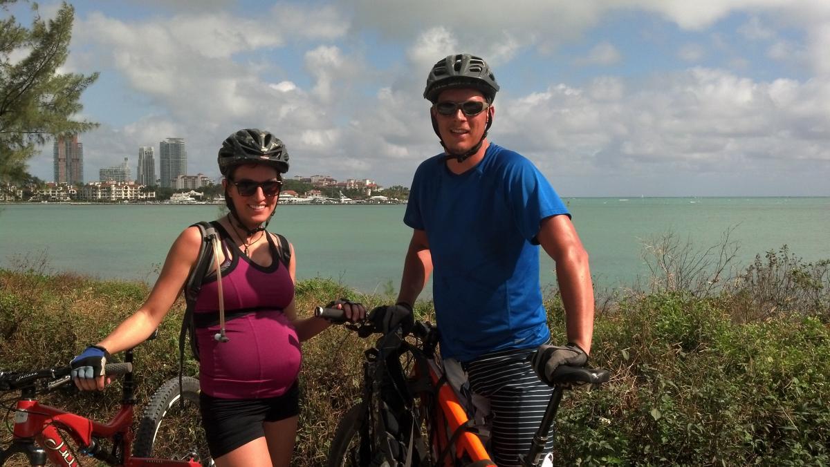 A pregnant woman and a man, both wearing helmets and sunglasses, are standing next to their bicycles along a coastal path. The background features calm turquoise water and a skyline with modern buildings under a partly cloudy sky. Virginia Key North Point mountain bike trail.