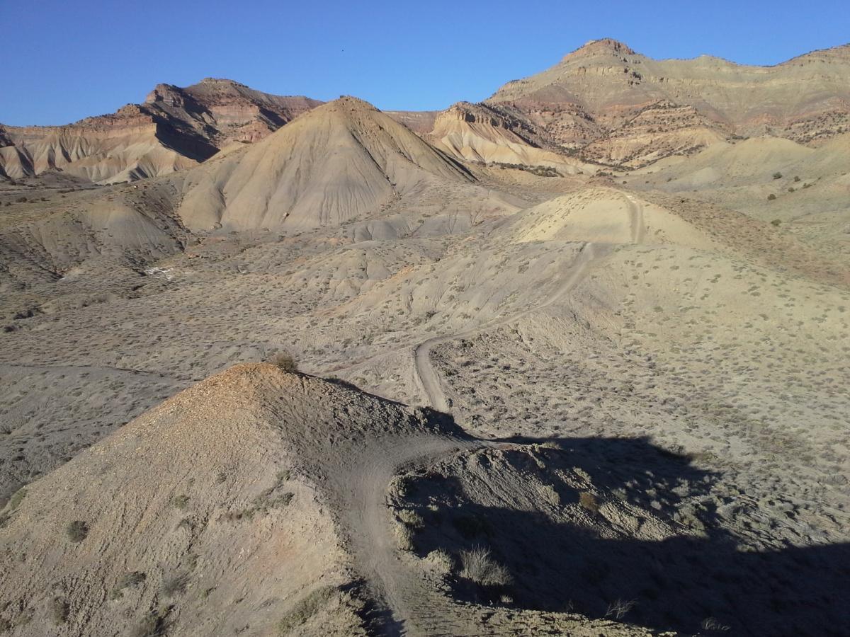 A panoramic view of a rugged desert landscape featuring rolling hills and rocky terrain under a clear blue sky. The ground is primarily dry and barren, with patches of sparse vegetation and a winding dirt path visible through the hills. The scene showcases the natural geological formations and contours typical of arid environments. 18 Road Trails / North Fruita Desert mountain bike trail.