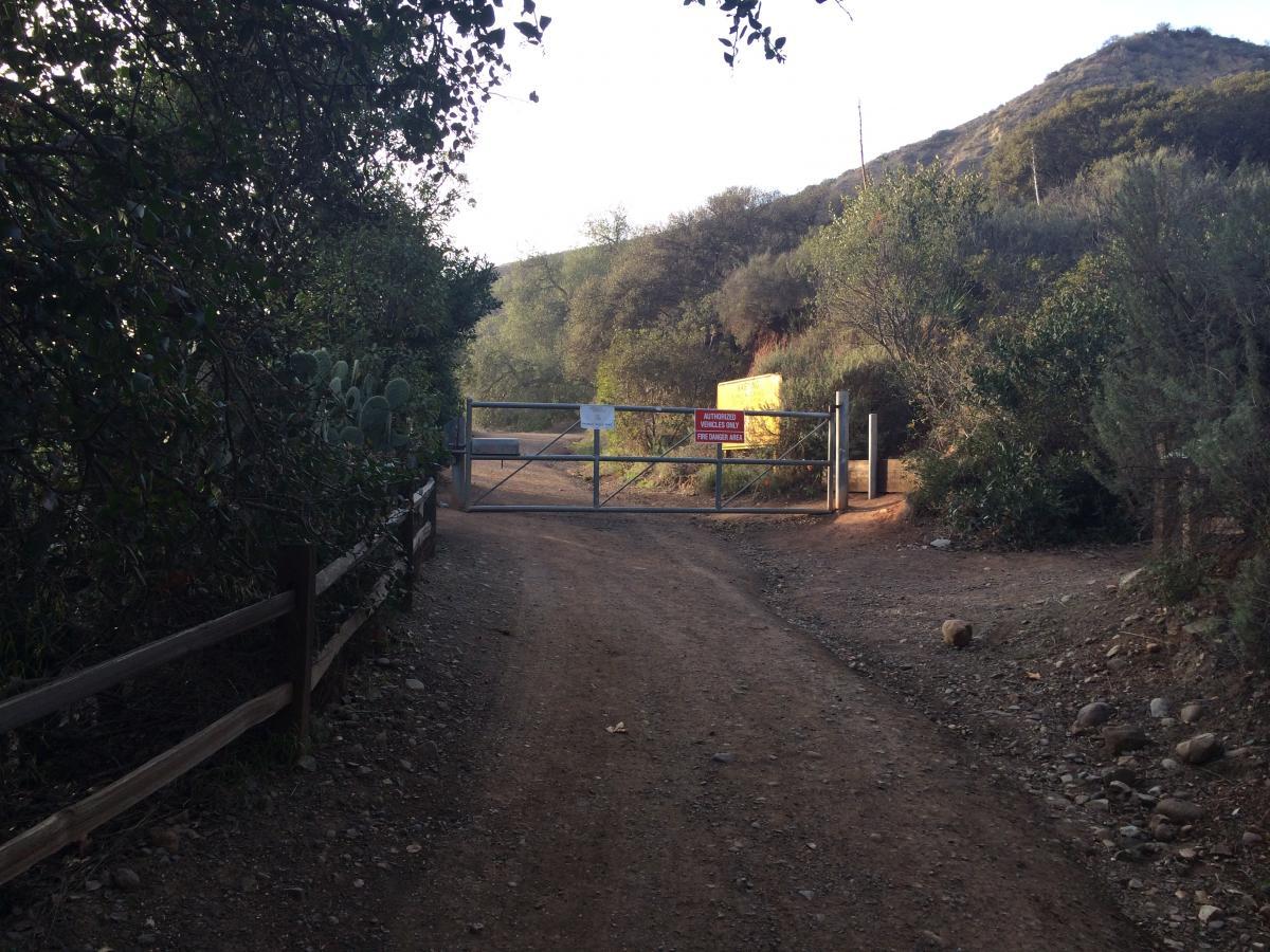 A dirt pathway leading to a metal gate surrounded by greenery, with a warning sign visible on the gate. The scene is set against a hillside in the background, suggesting a natural landscape. Harding Truck Trail mountain bike trail.