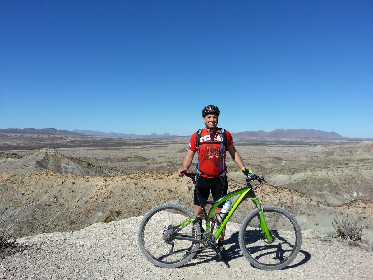 Niner Jet 9 RDO: A mountain biker wearing a red cycling jersey stands beside a green mountain bike on a rocky terrain, with a vast desert landscape and clear blue sky in the background.