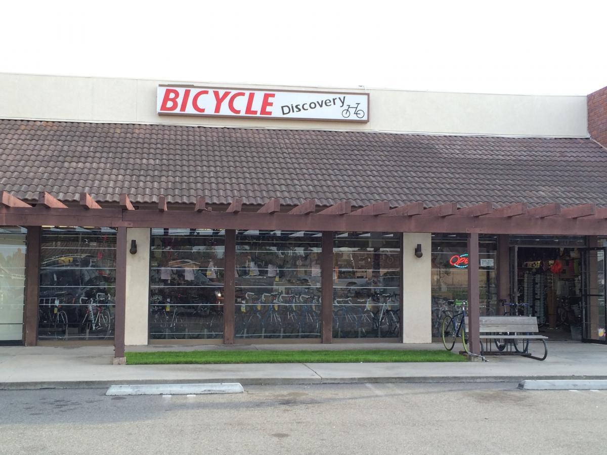 Front view of a bicycle store named "Bicycle Discovery," featuring a large window displaying various bicycles inside. The store has a brown tiled roof and wooden accents, with a bench in front and a neon "Open" sign visible through the entrance.
