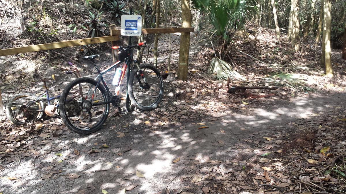 A mountain bike parked next to a trail surrounded by trees and foliage, with a wooden fence in the background. A sign indicating "Washing Station Entrance Intermediate" is visible nearby. The ground is covered in dirt and fallen leaves, suggesting an outdoor recreational area. Markham Park mountain bike trail.