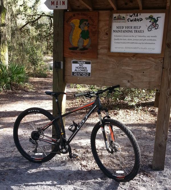 GT Karakoram: A mountain bike leaning against a trail sign with various information, including a notice about no horses allowed and a request for volunteers to help maintain the trails. The background features a wooded area with trees and foliage.