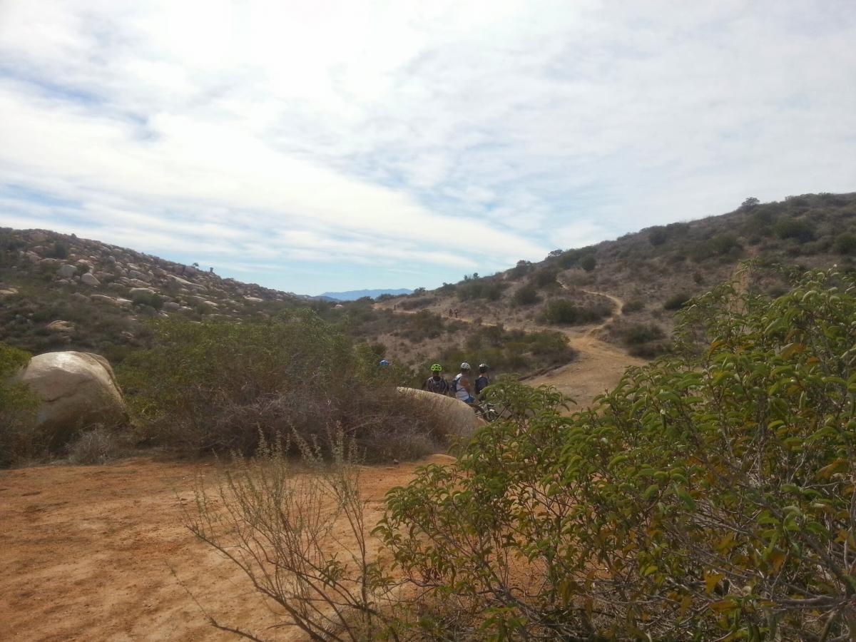 A scenic outdoor landscape featuring rolling hills and a dirt path, surrounded by shrubs and natural vegetation. Two cyclists are visible in the distance, riding along the trail, under a partly cloudy sky. Boulders and gentle slopes add to the tranquil, natural setting. Sycamore Canyon mountain bike trail.