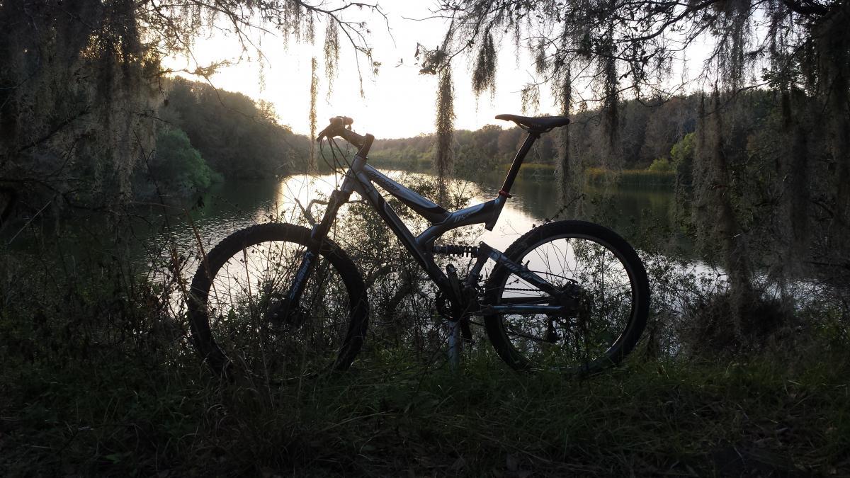 Giant Warp DS2: A mountain bike silhouetted against a peaceful river landscape at sunset, surrounded by greenery and hanging moss.